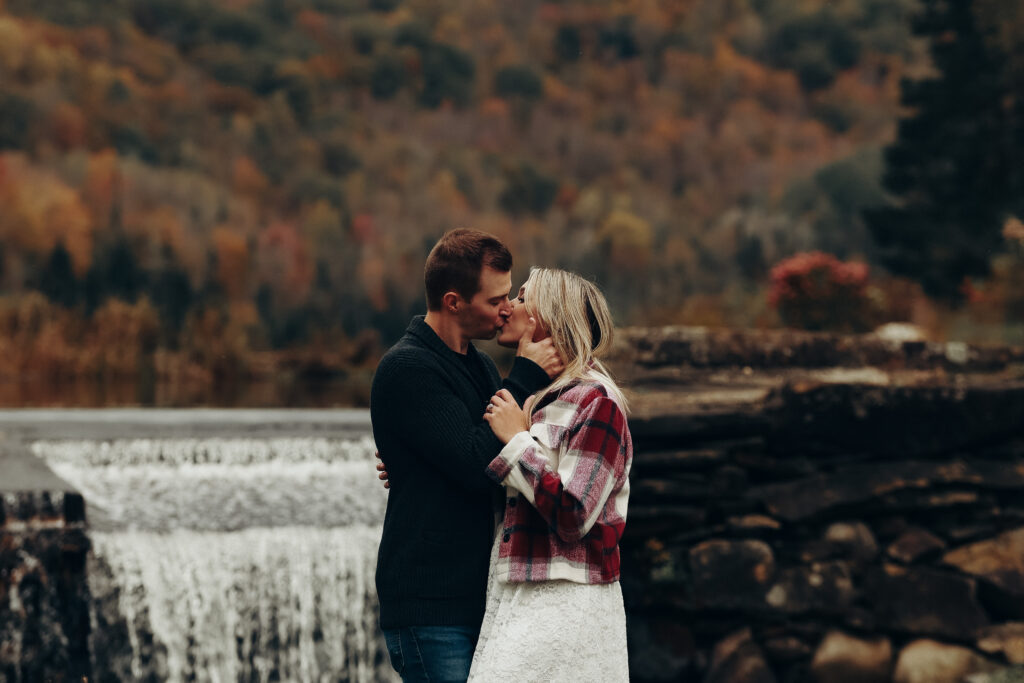 Couple kissing in front of a waterfall in Adamant, Vermont. The couple posed for their engagement photography session. Fall leaves have changed and the beautiful fall colors are showing in the phots. 